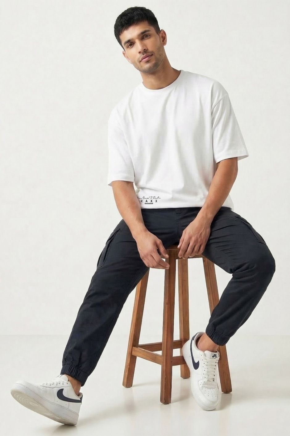 Man sitting on a wooden stool wearing a white t-shirt and black pants against a white background