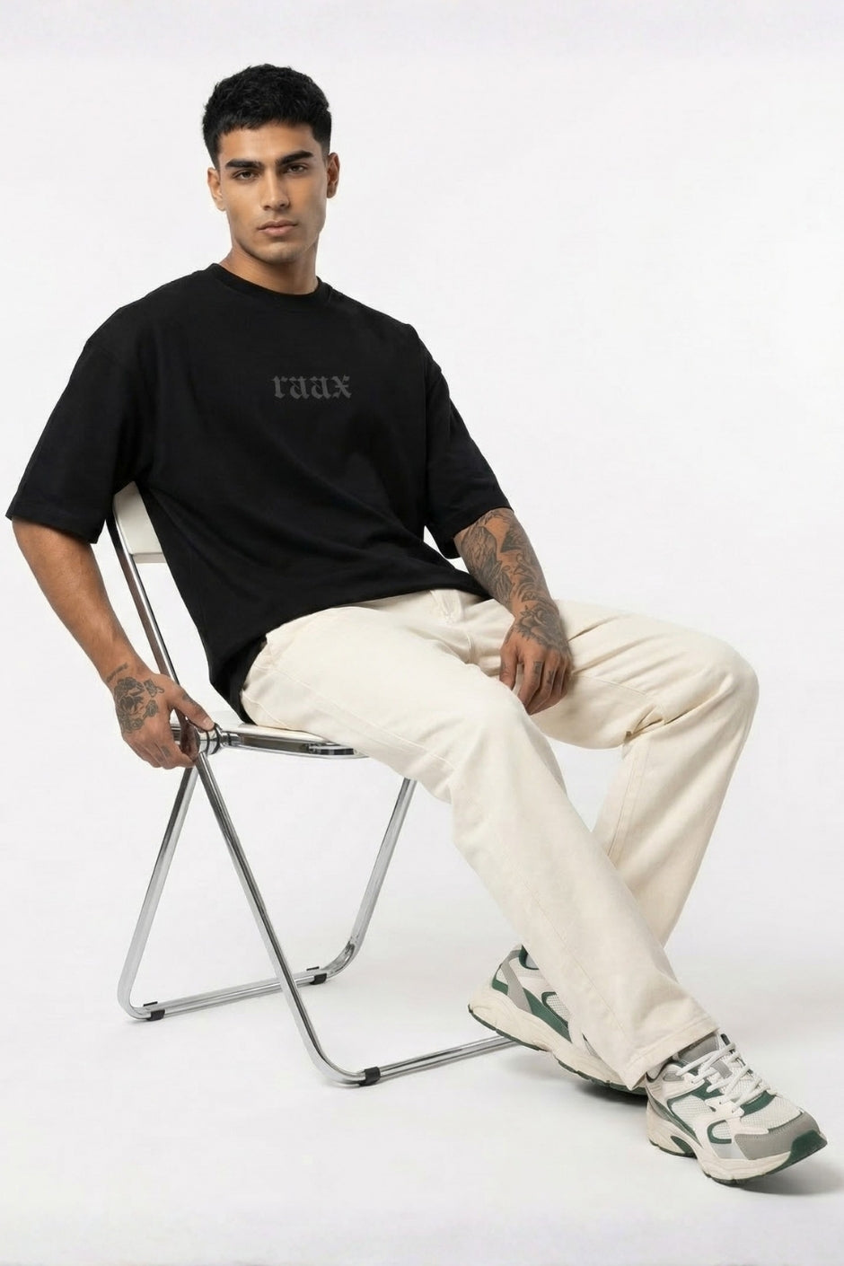 Man sitting on a chair wearing a black t-shirt with text, white pants, and sneakers on a white background