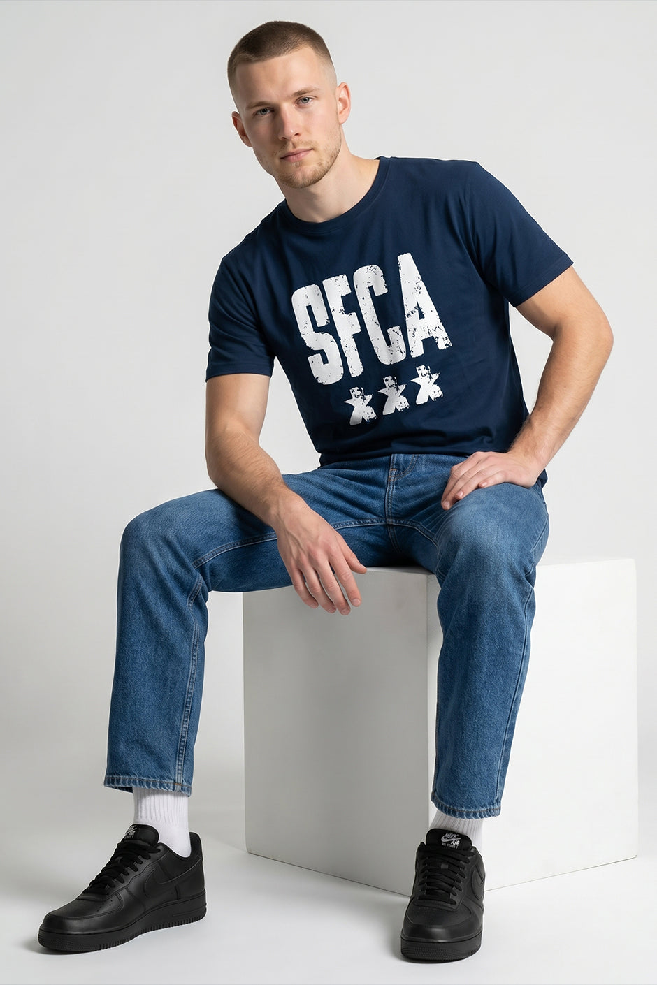 Man wearing a pastel blue t-shirt with 'SFCA' text and graphics, sitting on a white cube.