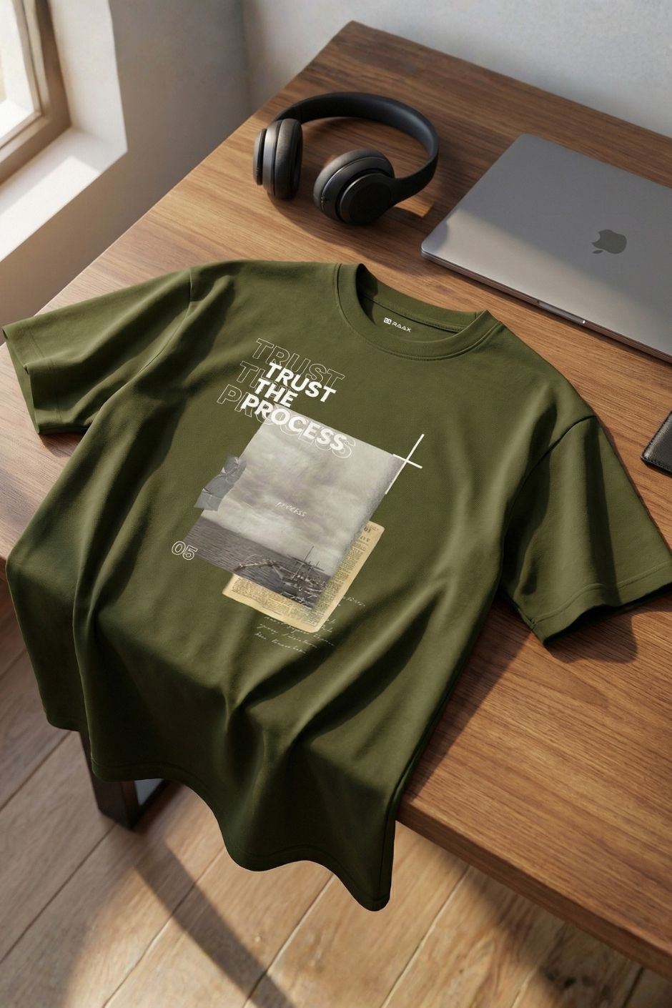 Olive t-shirt with text and graphic on a wooden table with headphones and a laptop.