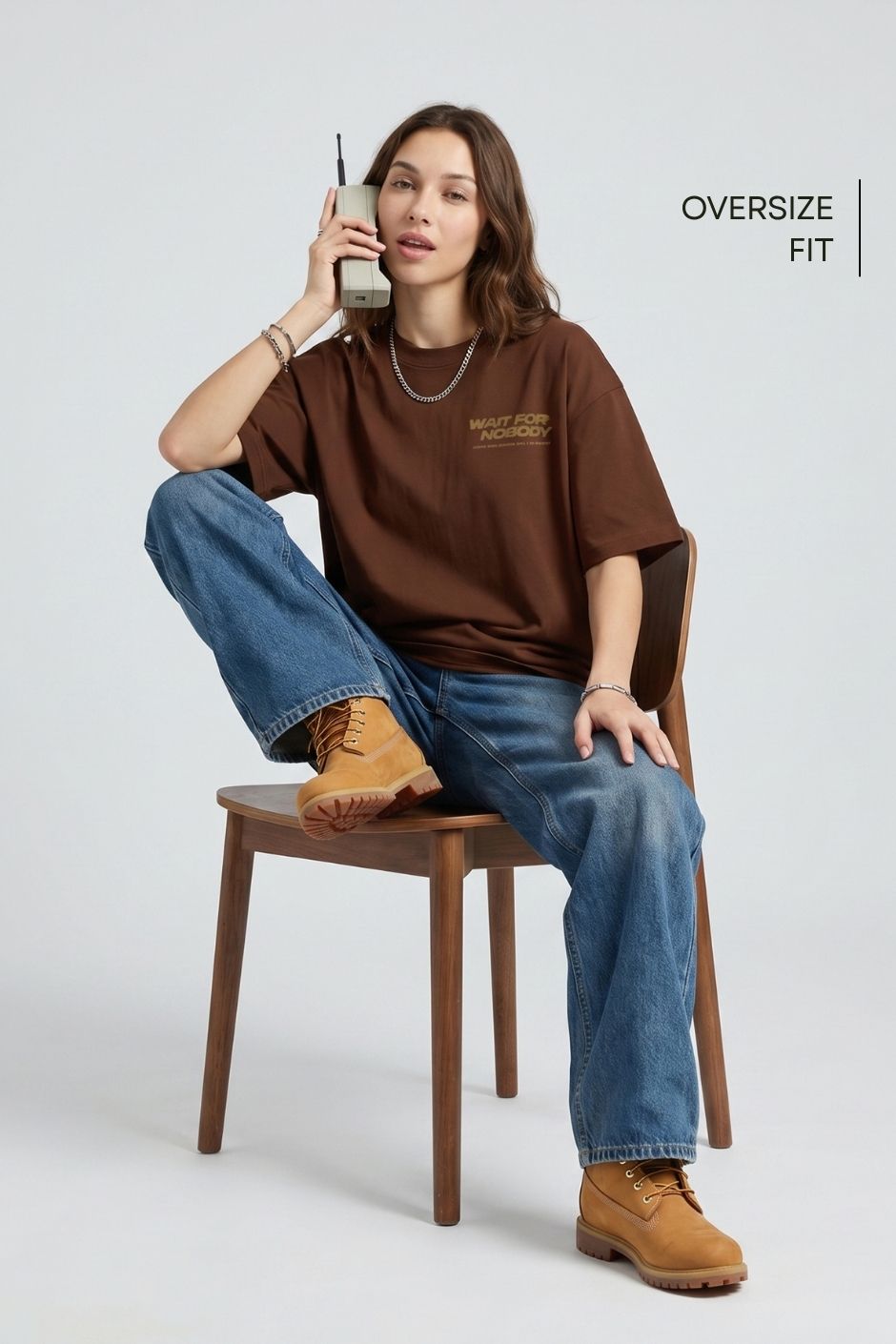 Person wearing an oversized brown t-shirt, blue jeans, and tan boots sitting on a wooden chair.