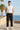 Man wearing a white sleeveless shirt and black cargo jogger standing by the ocean with palm trees in the background.