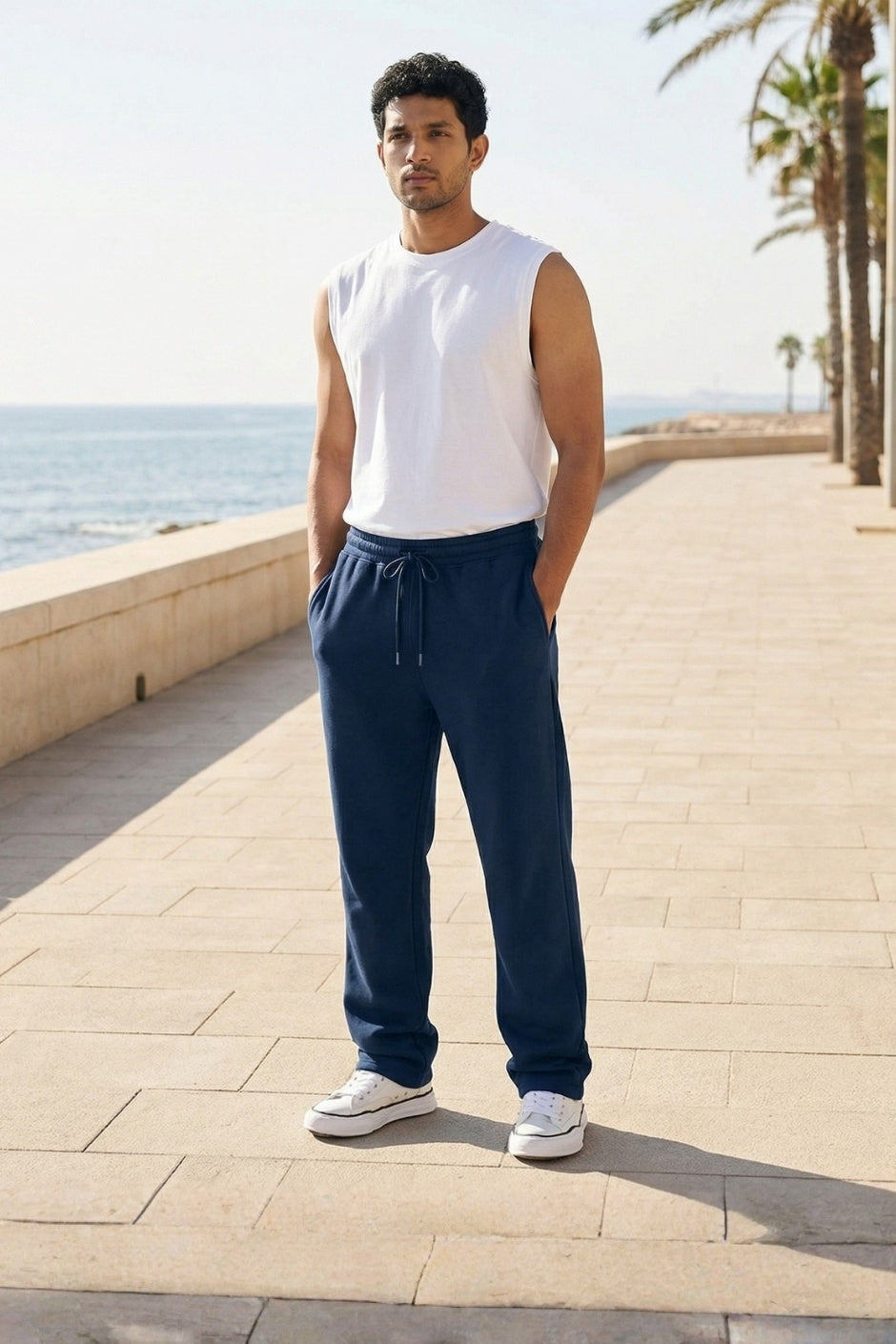 Man wearing a white sleeveless shirt and pastel blue jogger standing on a pathway by the sea.