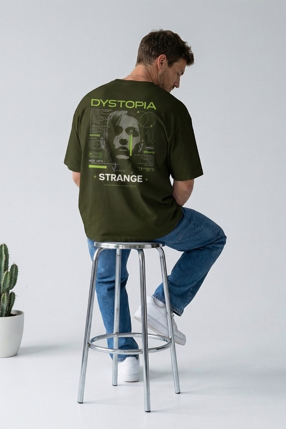 Person wearing a olive t-shirt with text and graphics, sitting on a stool against a plain background.