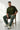 Man sitting on a wooden chair wearing a green 'DYSTOPIA' t-shirt against a white background