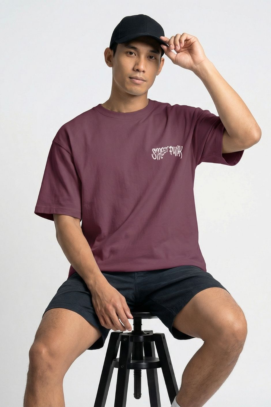 Person wearing a plum t-shirt with text and black shorts, sitting on a stool against a white background