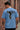 Man wearing a light blue t-shirt with a graphic design on the back, standing against a brick wall.