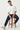 Man sitting on a wooden stool wearing a white t-shirt and black pants against a white background