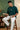 Man sitting on a stool reading a book and holding a mug in a room with green walls and a wooden shelf.