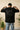 Person wearing a black 'RAAX' t-shirt with text on the back, standing indoors with plants in the background.