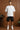 Man wearing a white t-shirt and black shorts standing against a wooden paneled wall.