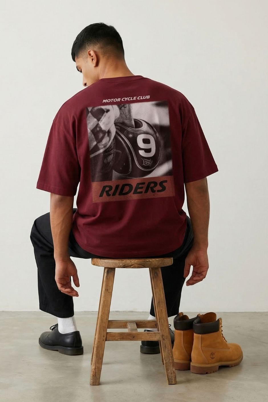 Person wearing a rust t-shirt with motorcycle graphic and text, sitting on a wooden stool.