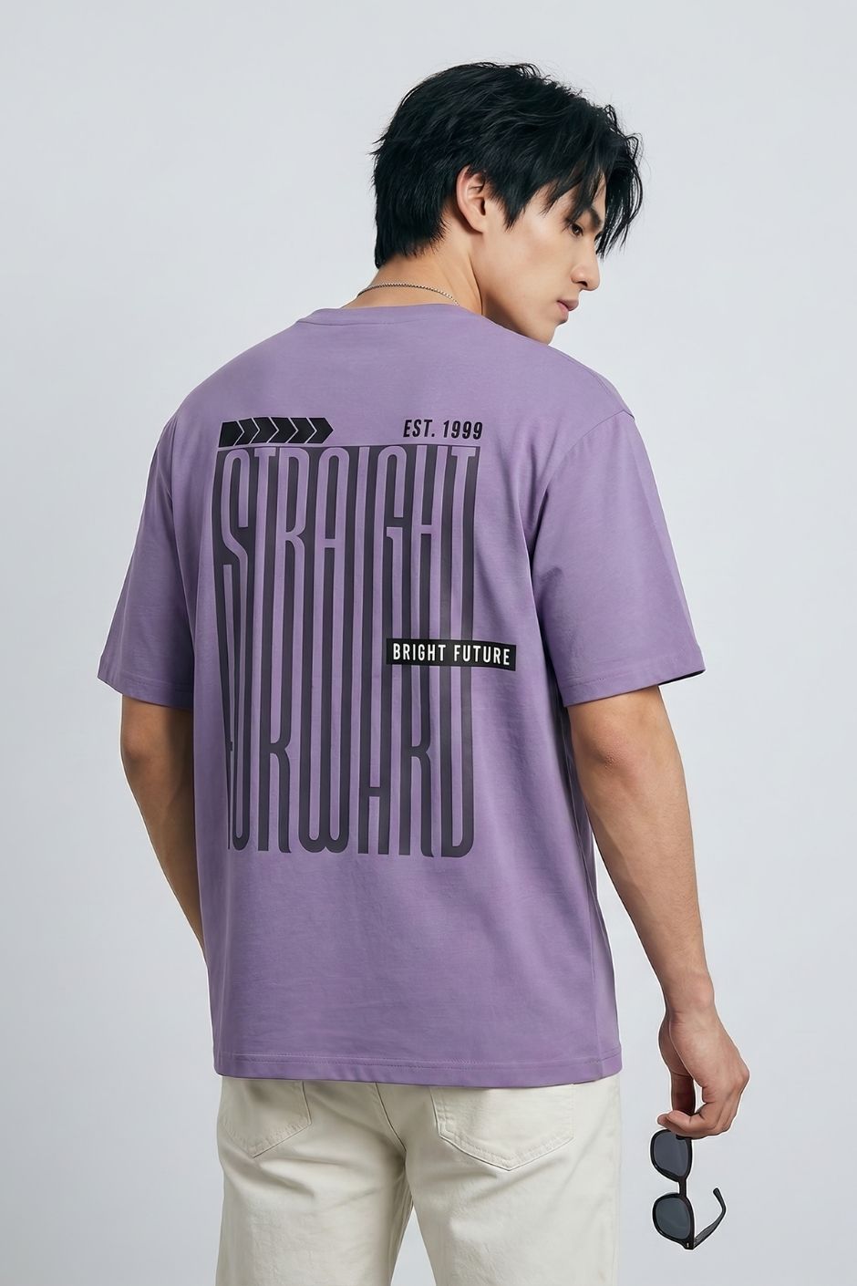 Person wearing a lilac t-shirt with text on the back, holding sunglasses, against a plain background