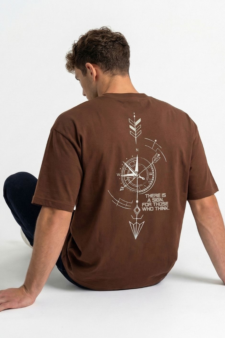 Person wearing a brown t-shirt with a compass design and text on the back, sitting on a white floor.