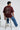 Man wearing a maroon red t-shirt with a geometric design on the back, sitting on a stool against a white background.