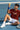 Person sitting on a tennis court holding a tennis ball, wearing a rust shirt and white shorts.