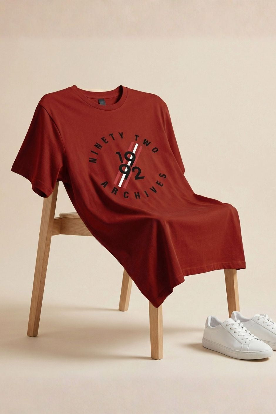 Rust t-shirt with 'Ninety Two Archives' design on a wooden chair with white sneakers beside it.