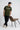 Man wearing a olive t-shirt with text standing next to a brown chair on a white background