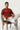 Man sitting on a chair wearing a rust t-shirt and beige pants against a white background