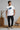 Man wearing a white t-shirt with a blue graphic and text, standing in a room with wooden furniture and decor.