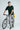 Man holding a bicycle wearing a bottle green t-shirt and gray pants on a light gray background