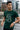 Man wearing a bottle green t-shirt with text and graphics, standing by a waterfront with buildings in the background.