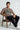 Man sitting on a chair wearing a chocolate t-shirt with graphic design and text.