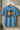 Light blue t-shirt with graphic design on a hanger against a neutral wall.