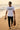 Man walking on a beach wearing a white t-shirt with text, holding sunglasses and a bag.