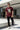 Man wearing a maroon red t-shirt with graphic design, black pants, and white sneakers standing in a modern outdoor setting.