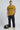 Man wearing a mustard yellow t-shirt with 'TRUST' text, black pants, and yellow shoes on a light gray background.