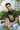 Man sitting on a bench wearing a olive t-shirt with text and headphones, surrounded by greenery.