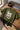 Olive t-shirt with text and graphic on a wooden table with headphones and a laptop.
