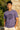 Man wearing a lilac t-shirt with text standing against a stone wall.