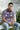 Man sitting on a bench wearing a lilac t-shirt with text, holding headphones and a phone.