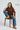 Person wearing an oversized brown t-shirt, blue jeans, and tan boots sitting on a wooden chair.